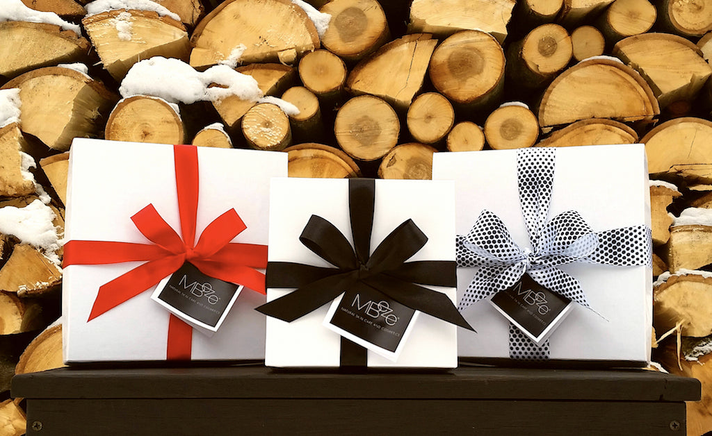 Three MBeze gift boxes in front of a stack of chopped wood and snow.