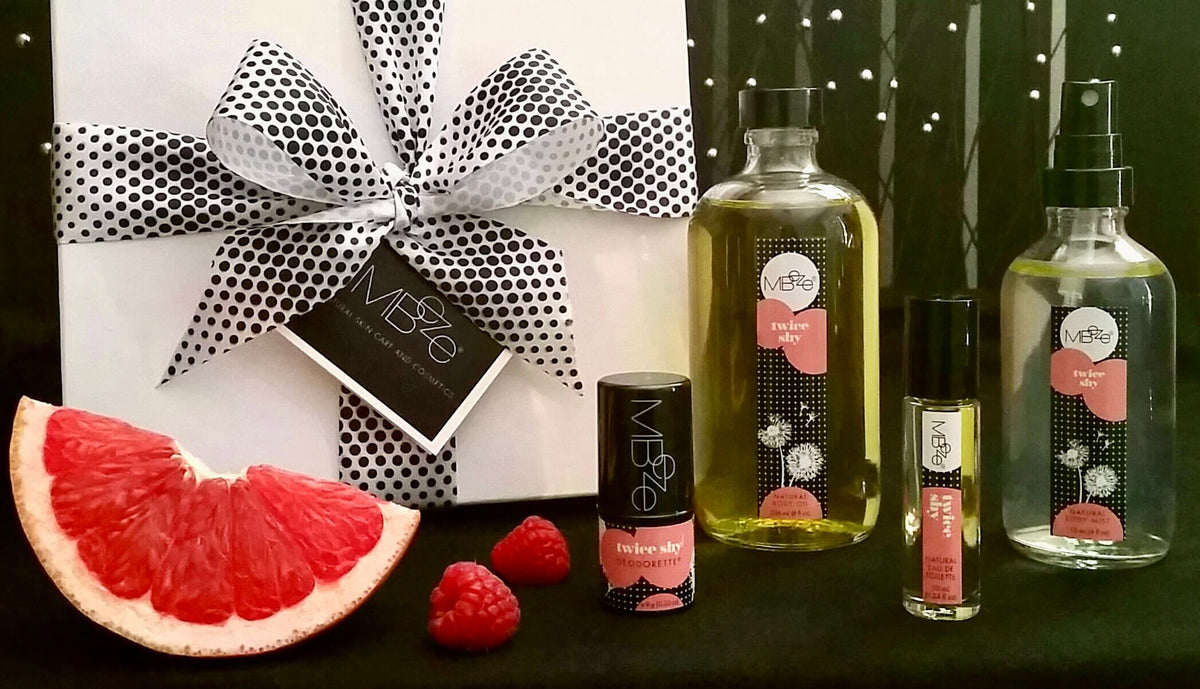 MBeze Twice Shy Collection including the gift box, Deodorette, Body Oil, Perfume, and Body Mist with a grapefruit slice and raspberries.