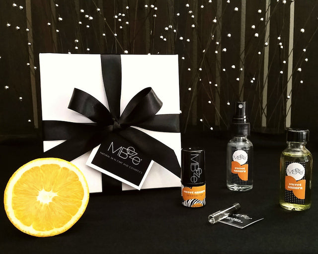 MBeze Sweet Sanura Mini Gift Set with gift box. Including Deodorette, Body Mist Traveler, Body Oil Traveler, Perfume Sample, with an orange slice in front.